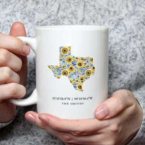 May include: White ceramic mug featuring a floral Texas state design with sunflowers and blue flowers. The mug includes the coordinates 32°35'58.9"N | 99°39'50.4"W and the name "THE SMITHS".