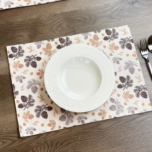 May include: A white ceramic dinner plate with a decorative rim sits on a beige placemat with a brown and orange pine cone pattern. A fork and spoon are on the table to the right of the plate.