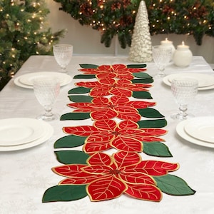 May include: A red and green poinsettia table runner with gold accents. The runner is made of fabric and features a repeating pattern of poinsettia flowers and leaves.