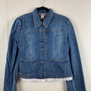 Puede incluir: Una chaqueta vaquera azul con ribete de encaje blanco alrededor del dobladillo y las mangas. La chaqueta tiene cierre de botones y dos bolsillos delanteros. La etiqueta de la chaqueta dice "CLIO".