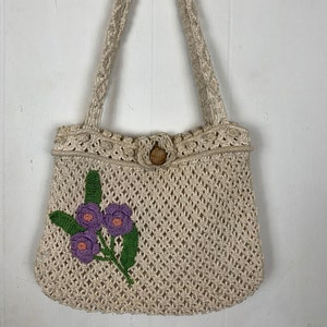 May include: A crocheted tote bag with a floral design. The bag is made of natural fibers and has a wooden button closure. The floral design features purple flowers and green leaves.