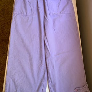 May include: Lavender-colored cropped pants with an elastic waistband and two front pockets. The pants have a relaxed fit and feature button details at the hem. The fabric appears to be a lightweight cotton or blend.