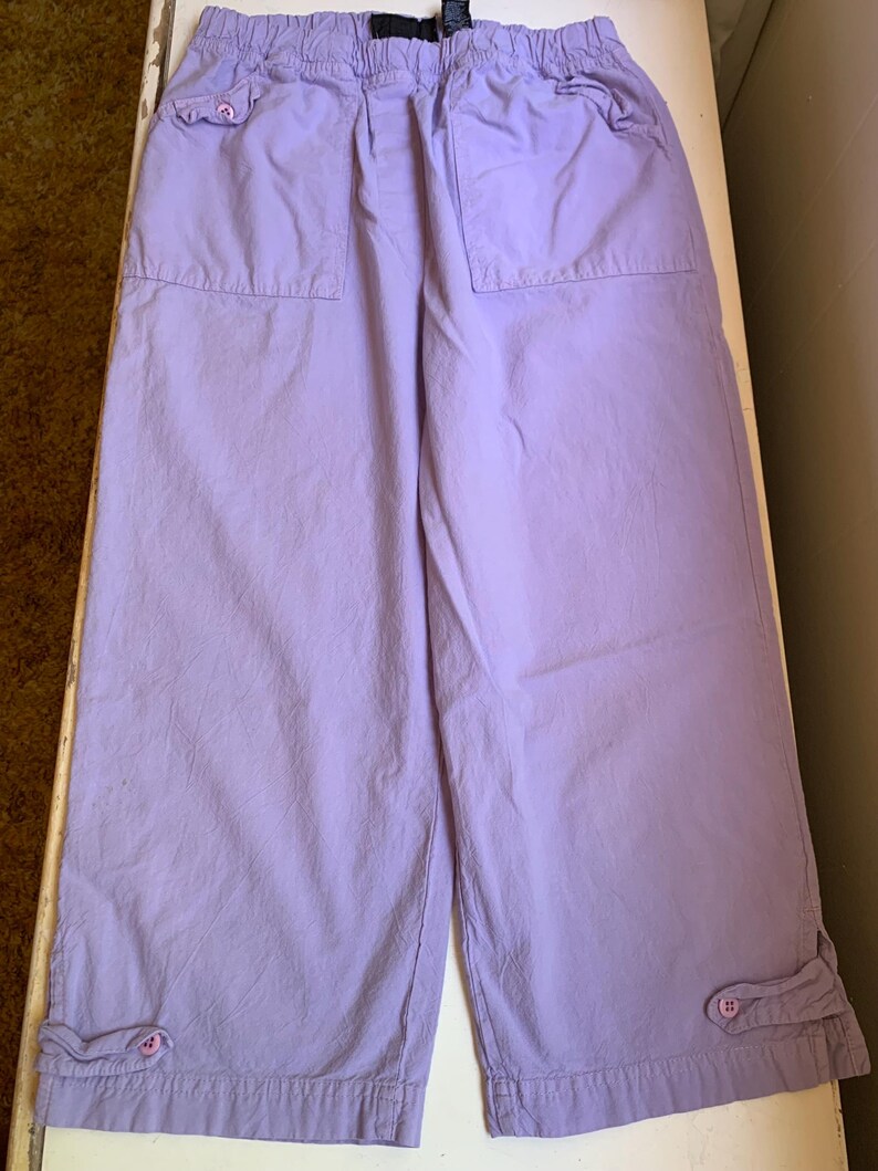 May include: Lavender-colored cropped pants with an elastic waistband and two front pockets. The pants have a relaxed fit and feature button details at the hem. The fabric appears to be a lightweight cotton or blend.