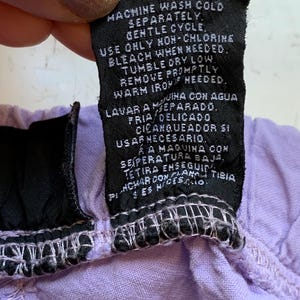 May include: A close-up of a black clothing tag with white text, held against a light purple fabric. The tag provides washing instructions, including machine wash cold, gentle cycle, and tumble dry low.