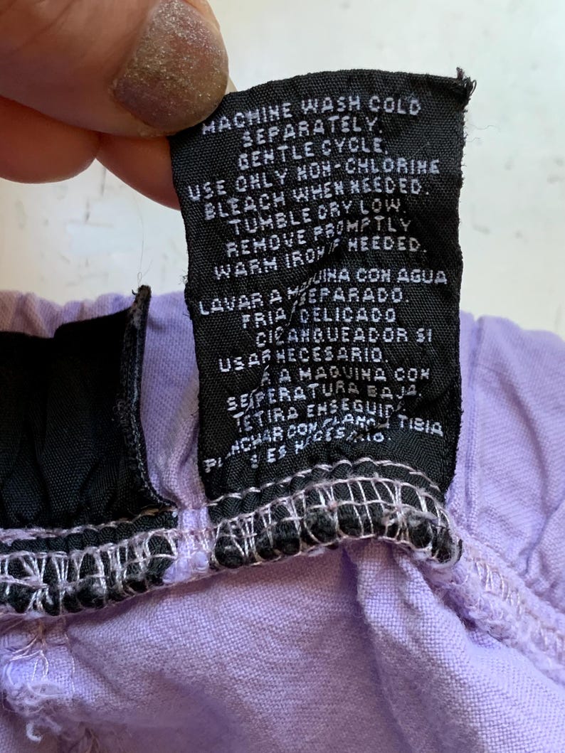 May include: A close-up of a black clothing tag with white text, held against a light purple fabric. The tag provides washing instructions, including machine wash cold, gentle cycle, and tumble dry low.