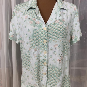 May include: A short-sleeved button-down shirt with a light blue background and a floral pattern in shades of green and pink. The shirt has a V-neck collar and is displayed on a mannequin.
