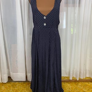 May include: A navy blue sleeveless maxi dress with a white polka dot pattern. The dress features a V-neckline, two white buttons, and a flowing skirt. The dress is displayed on a mannequin.