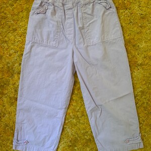 May include: Light purple capri pants with an elastic waistband and two front pockets. The pants have a relaxed fit and feature button details at the cuffs. The fabric appears to be a lightweight cotton blend, suitable for casual wear.