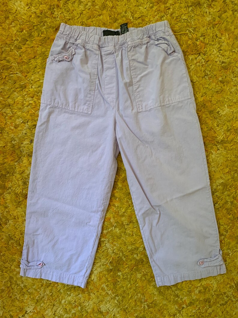May include: Light purple capri pants with an elastic waistband and two front pockets. The pants have a relaxed fit and feature button details at the cuffs. The fabric appears to be a lightweight cotton blend, suitable for casual wear.
