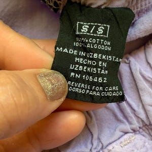 May include: Close-up of a black clothing tag with white text. The tag reads "S/S", "100% COTTON", and "MADE IN UZBEKISTAN". The tag is held against a light purple fabric. The tag also includes care instructions.