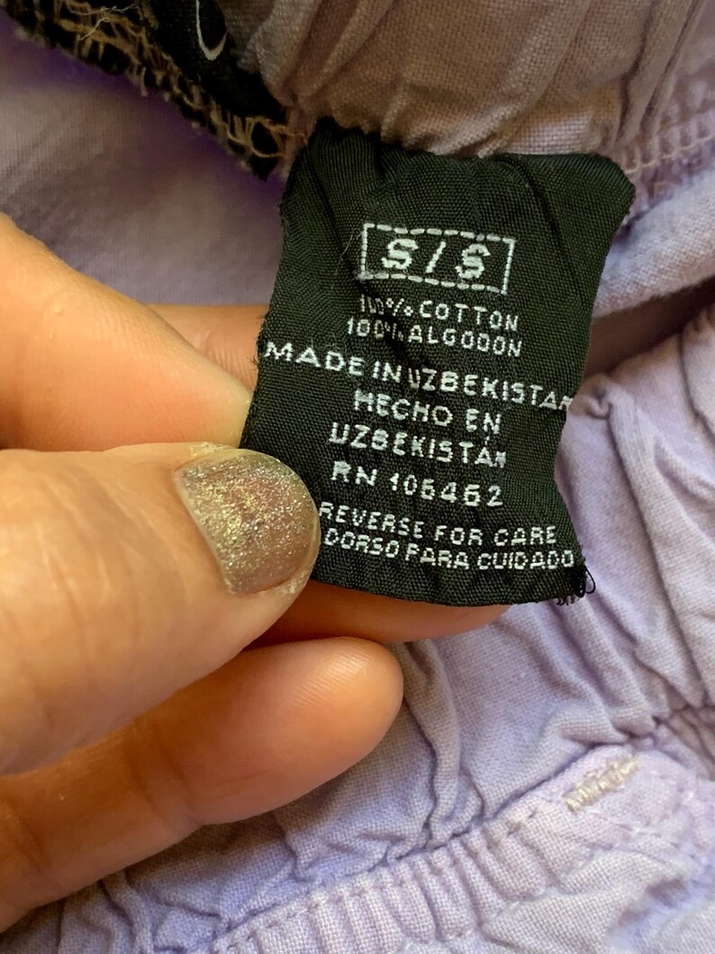 May include: Close-up of a black clothing tag with white text. The tag reads "S/S", "100% COTTON", and "MADE IN UZBEKISTAN". The tag is held against a light purple fabric. The tag also includes care instructions.