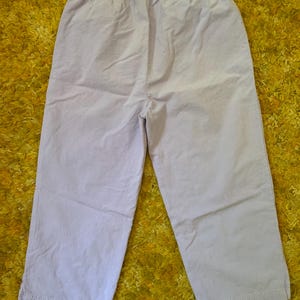 May include: Light purple cropped pants with an elastic waistband. The pants are made of a lightweight fabric and have a relaxed fit. The pants are shown on a yellow textured surface.
