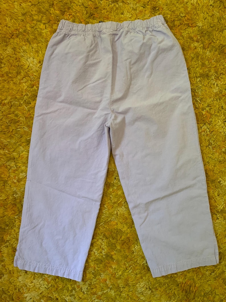 May include: Light purple cropped pants with an elastic waistband. The pants are made of a lightweight fabric and have a relaxed fit. The pants are shown on a yellow textured surface.