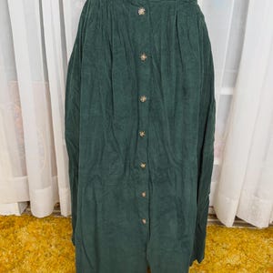 May include: A dark green corduroy skirt with a button-down front. The skirt has a high waist and a pleated design, with buttons down the center. The fabric appears soft and has a slight sheen. The skirt is a midi length.