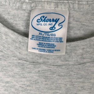 May include: Gray t-shirt tag with the text "Sherry MFG. CO. INC." in a white circle. The tag also includes the text "XL/TG/EG 98% Cotton Preshrunk/2% Other Fiber 98% Cotton Pre-Retreci/2% Autre Fibre 98% Algodon Pre-Encogido/2% Otra Fibra Made in Honduras Fabrique au Honduras Hecho en Honduras Care on Reverse/Soins au Revers Cuidado al Dorso"