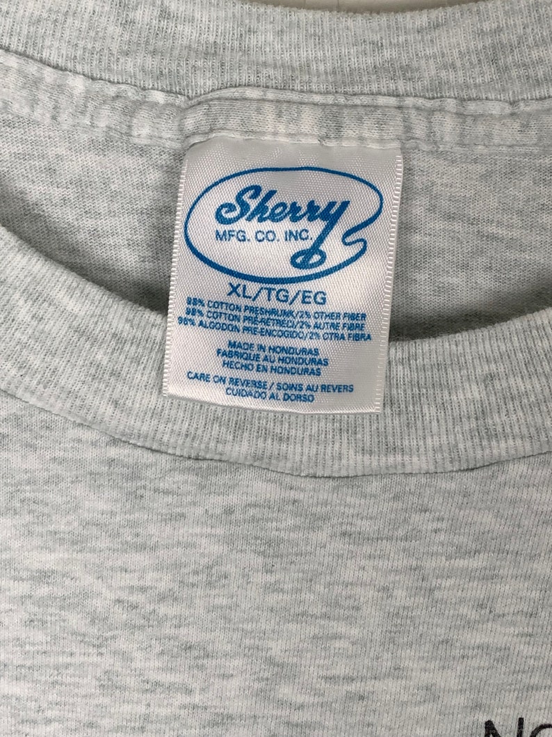 May include: Gray t-shirt tag with the text "Sherry MFG. CO. INC." in a white circle. The tag also includes the text "XL/TG/EG 98% Cotton Preshrunk/2% Other Fiber 98% Cotton Pre-Retreci/2% Autre Fibre 98% Algodon Pre-Encogido/2% Otra Fibra Made in Honduras Fabrique au Honduras Hecho en Honduras Care on Reverse/Soins au Revers Cuidado al Dorso"