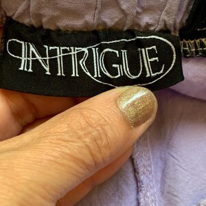 May include: Close-up of a black label with the word "INTRIGUE" in white script. The label is sewn onto a lavender fabric. The fabric is gathered at the top and has a textured appearance.