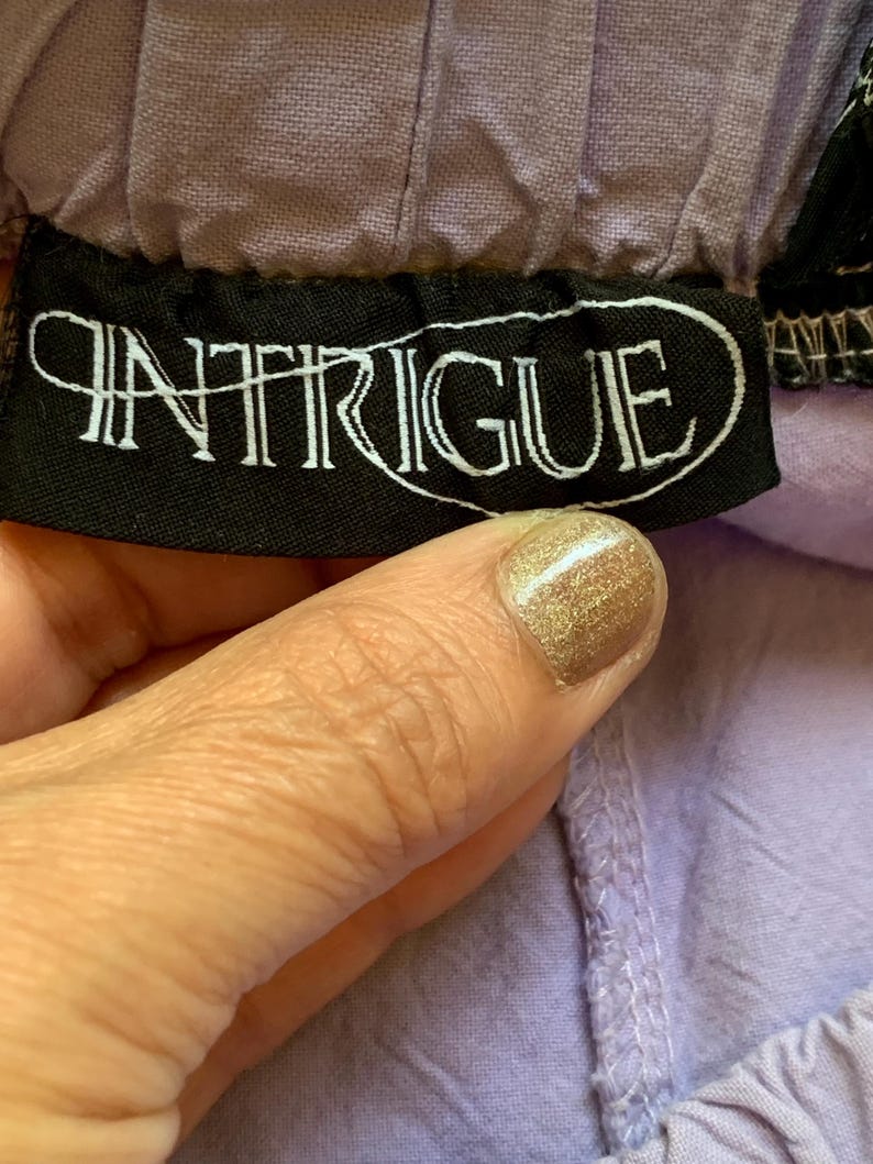 May include: Close-up of a black label with the word "INTRIGUE" in white script. The label is sewn onto a lavender fabric. The fabric is gathered at the top and has a textured appearance.