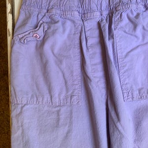 May include: Close-up of lavender-colored pants with an elastic waistband and two large pockets. One pocket has a decorative flap with a pink button. The fabric has a slightly wrinkled texture.