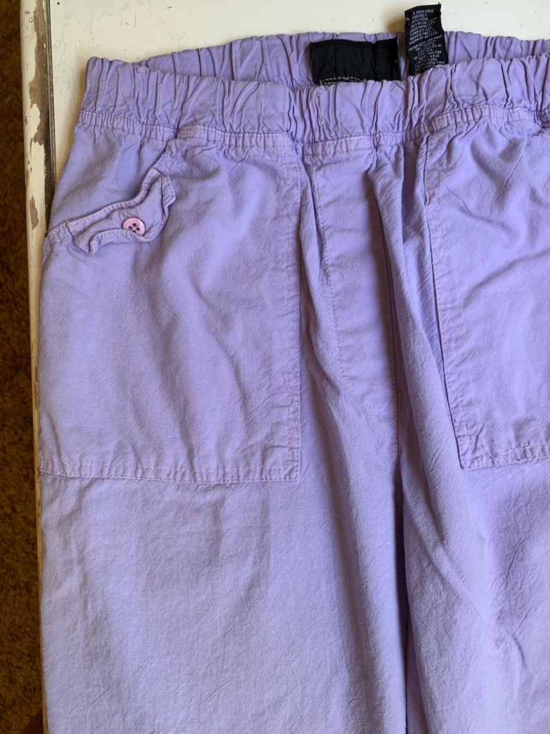 May include: Close-up of lavender-colored pants with an elastic waistband and two large pockets. One pocket has a decorative flap with a pink button. The fabric has a slightly wrinkled texture.