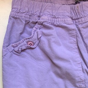 May include: Close-up of lavender-colored shorts with an elastic waistband. The shorts feature a ruffled pocket with a matching button. The fabric has a slightly wrinkled texture.