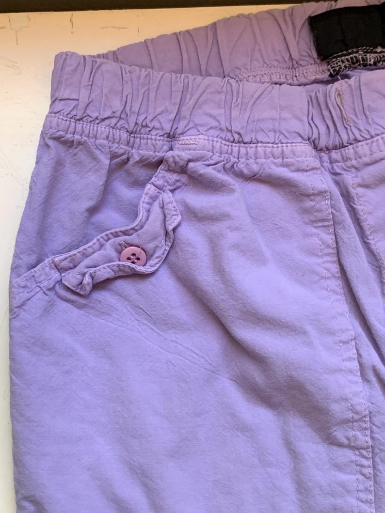 May include: Close-up of lavender-colored shorts with an elastic waistband. The shorts feature a ruffled pocket with a matching button. The fabric has a slightly wrinkled texture.