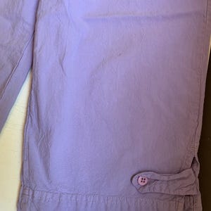 May include: Close-up of lavender-colored capri pants. The fabric is slightly wrinkled, with a button detail at the hem. The pants appear to be made of a lightweight material, suitable for warm weather. The stitching is visible along the hem.