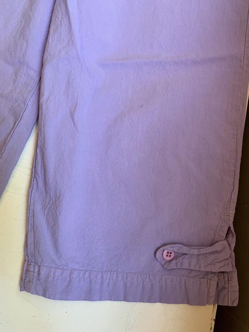 May include: Close-up of lavender-colored capri pants. The fabric is slightly wrinkled, with a button detail at the hem. The pants appear to be made of a lightweight material, suitable for warm weather. The stitching is visible along the hem.