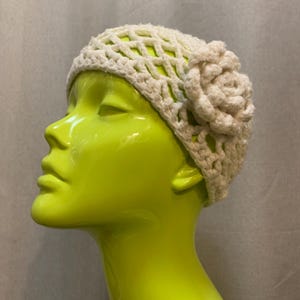 May include: A white crocheted hat with a large flower detail on the side. The hat is made with a delicate open stitch pattern.