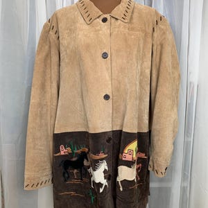 May include: Tan suede jacket with a dark brown bottom panel featuring embroidered horses and desert scenery. The jacket has a button-up front, notched collar, and decorative stitching along the collar and cuffs. A unique, western-themed outerwear piece.