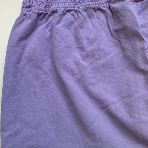 May include: Close-up of lavender-colored shorts with an elastic waistband. The fabric has a slightly wrinkled texture, suggesting a casual style. The shorts are a solid color, with no visible patterns or embellishments.