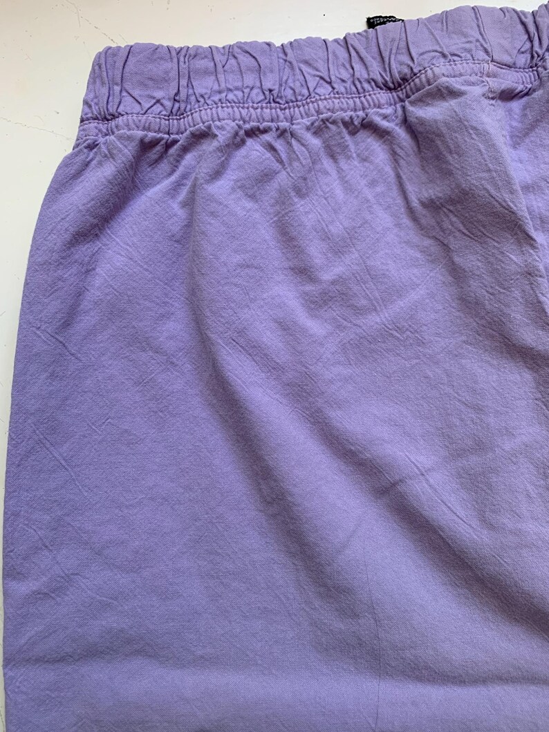 May include: Close-up of lavender-colored shorts with an elastic waistband. The fabric has a slightly wrinkled texture, suggesting a casual style. The shorts are a solid color, with no visible patterns or embellishments.