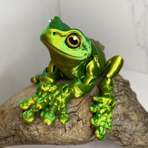 May include: A green 3D printed frog figurine with a textured surface and large, realistic eyes. The frog is sitting on a piece of driftwood.