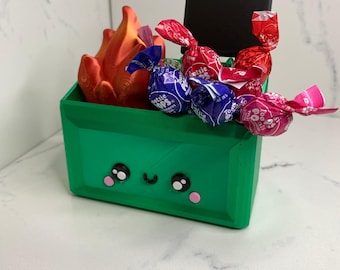 May include: A green, cartoon-style trash can with a happy face, filled with red, pink, and purple candy. The candy is wrapped in clear plastic with the words "Tootsie Pops" printed on it. There is a small black sign on top of the trash can.