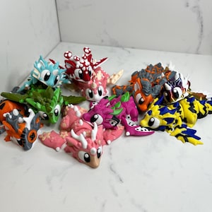May include: A collection of small, articulated dragon figurines in various colors including red, pink, green, blue, orange, yellow, and purple. Each dragon features large white eyes, detailed scales, and horns, with some displaying floral patterns.