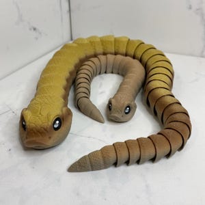 May include: Two articulated snake toys. The larger snake is yellow and brown, while the smaller one is tan. Both have detailed scales and large, reflective eyes. The snakes are posed in a curved position, suggesting movement. The toys are made of a textured material.