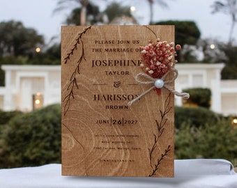 Pode incluir: Um convite de casamento de madeira com um fundo marrom rústico. O convite apresenta os nomes Josephine Taylor e Harisson Brown, a data 26 de junho de 2027 e o local do casamento. O convite é decorado com um pequeno buquê de flores secas e uma pérola.