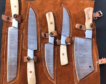 May include: A set of five knives with cream-colored handles and patterned blades, displayed in a brown leather roll. Each knife is secured with a leather strap and brass hardware. The blades have a unique, textured design.