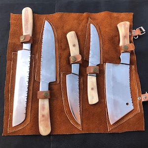 May include: A collection of five stainless steel knives with cream-coloured handles and brown leather straps, presented in a brown leather knife roll. The blades vary in size and shape, including a cleaver and serrated knives.