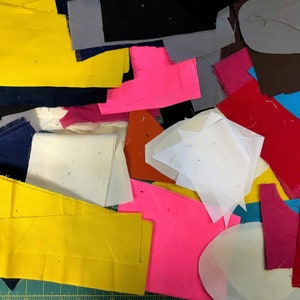 May include: A pile of assorted fabric offcuts in a range of colours and forms. Included are pieces of bright yellow, hot pink, red, black, and blue fabrics. Some are rectangular, others are irregular. The fabrics appear to be cotton or a similar material.