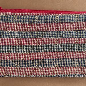 May include: A quilted pouch with a red zipper. The pouch is made of fabric with a checkered pattern in alternating rows of navy blue and red. The stitching is visible, creating a textured appearance.