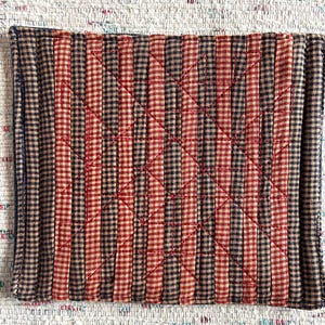 May include: Rectangular quilted placemat with a patchwork design. The fabric features a pattern of alternating vertical stripes in red, black, and tan gingham. Red thread creates a diamond pattern across the surface.