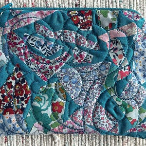 May include: A rectangular zippered pouch featuring a quilted patchwork design. The surface consists of solid teal fabric sections interspersed with various floral-patterned fabric pieces in shades of pink, red, blue, and green, all joined by visible stitching.