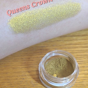 Puede incluir: Un peque&ntilde;o tarro de sombra de ojos de color amarillo dorado con el texto "Queens Crown" escrito en rosa sobre un fondo blanco.