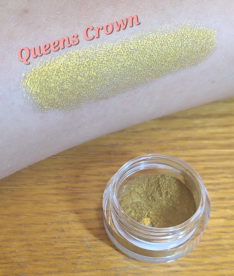 Puede incluir: Un peque&ntilde;o tarro de sombra de ojos de color amarillo dorado con el texto "Queens Crown" escrito en rosa sobre un fondo blanco.