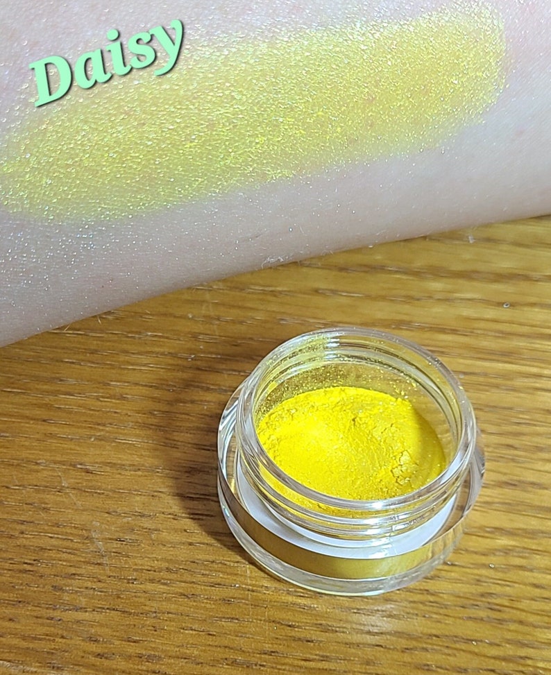 Puede incluir: Un peque&ntilde;o tarro de pigmento de sombra de ojos amarillo brillante etiquetado como "Daisy". El pigmento se muestra aplicado en la piel, revelando un color amarillo vibrante con un brillo sutil.