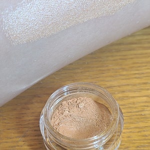 Puede incluir: Un peque&ntilde;o tarro de sombra de ojos dorada suelta con la palabra "Angora" escrita en amarillo sobre un fondo blanco. La sombra de ojos se muestra aplicada en la piel.