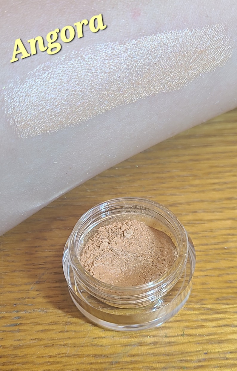 Puede incluir: Un peque&ntilde;o tarro de sombra de ojos dorada suelta con la palabra "Angora" escrita en amarillo sobre un fondo blanco. La sombra de ojos se muestra aplicada en la piel.