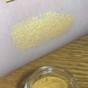 Puede incluir: Un peque&ntilde;o tarro de sombra de ojos de color amarillo dorado con la palabra "Honey" escrita en letras amarillas sobre una superficie marr&oacute;n claro.