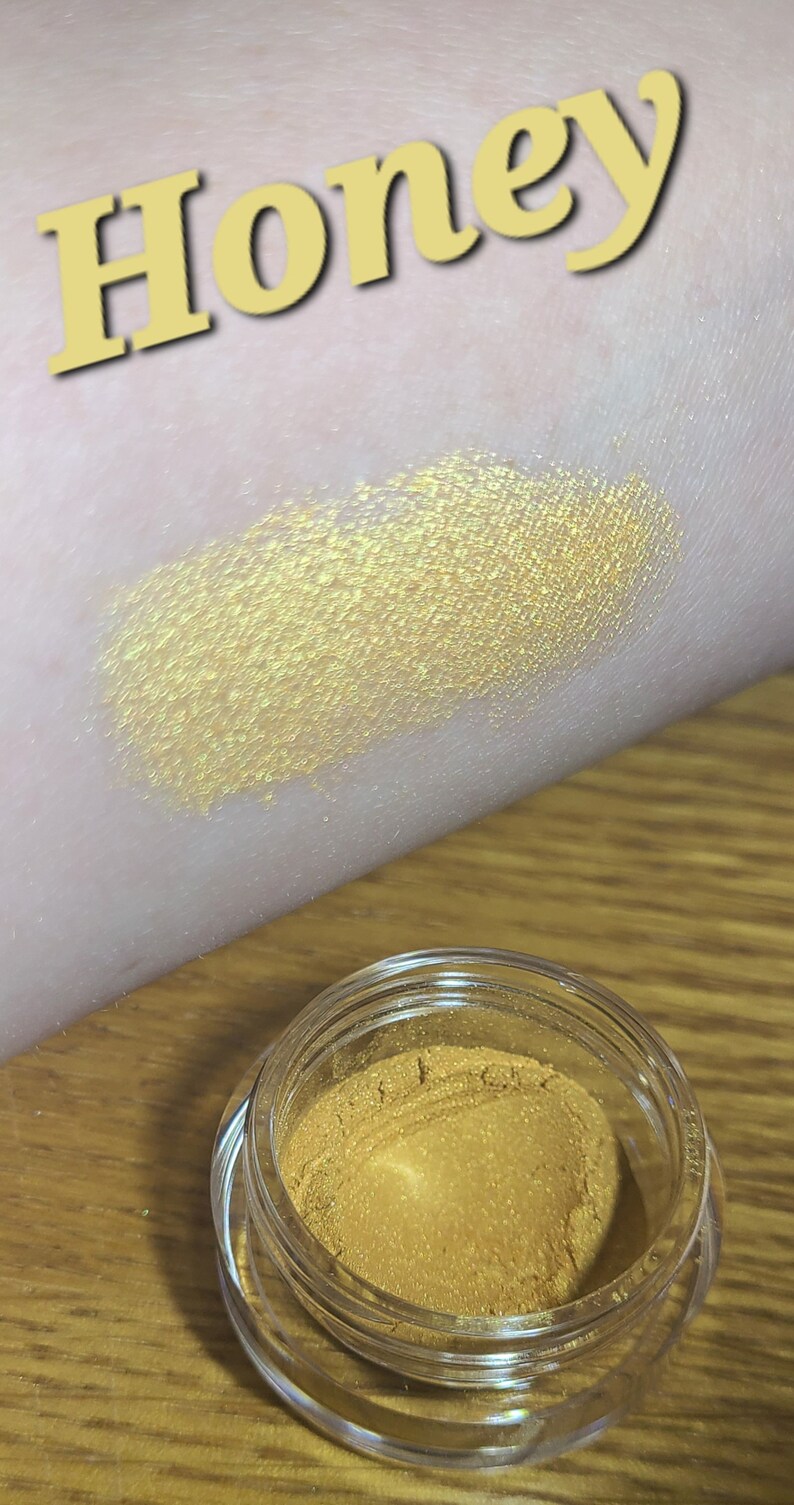 Puede incluir: Un peque&ntilde;o tarro de sombra de ojos de color amarillo dorado con la palabra "Honey" escrita en letras amarillas sobre una superficie marr&oacute;n claro.